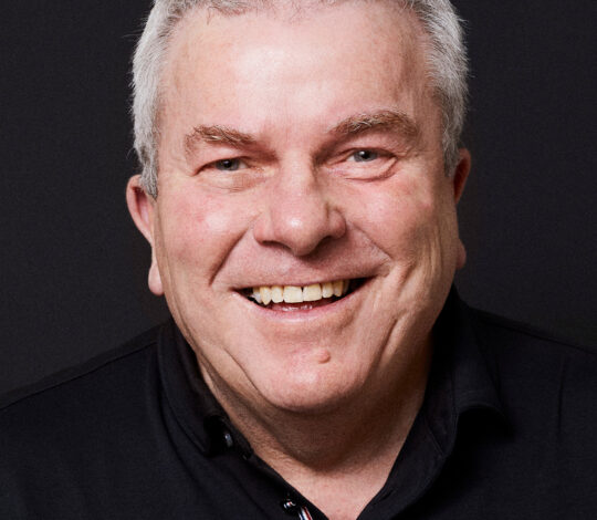 A smiling older man with short white hair and a black collared shirt against a dark background.