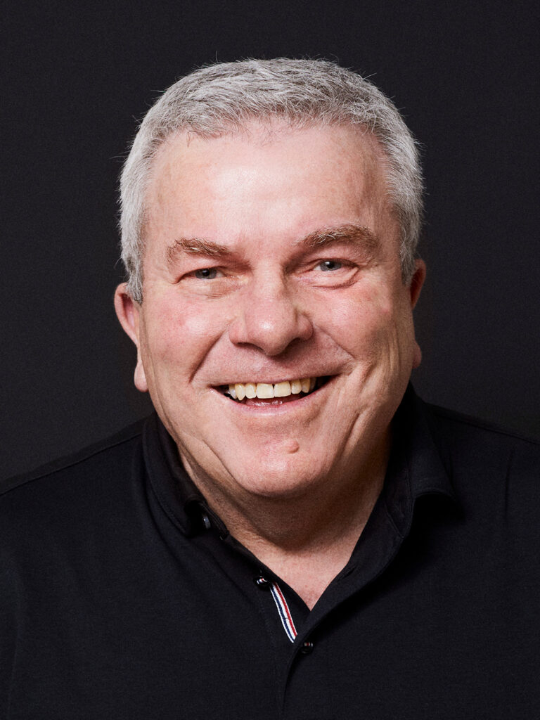 A smiling older man with short white hair and a black collared shirt against a dark background.