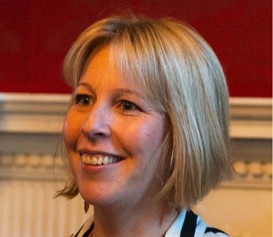 A woman with blonde hair and a striped shirt smiles while looking slightly to the side.