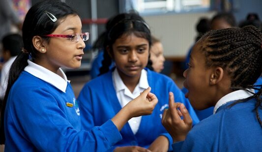 Four students in blue school uniforms engage in a conversation in a classroom. One student gestures with her hand while speaking to another, who appears to be listening attentively.