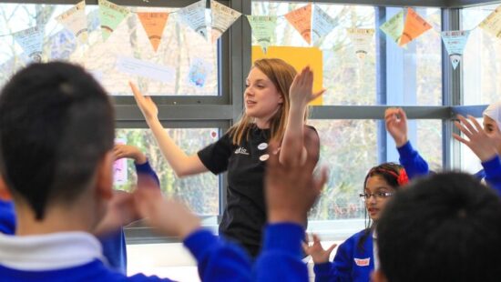 A female teacher stands in a classroom, engaging with students who are raising their hands. The students wear blue uniforms and colorful flags are hung in the background.