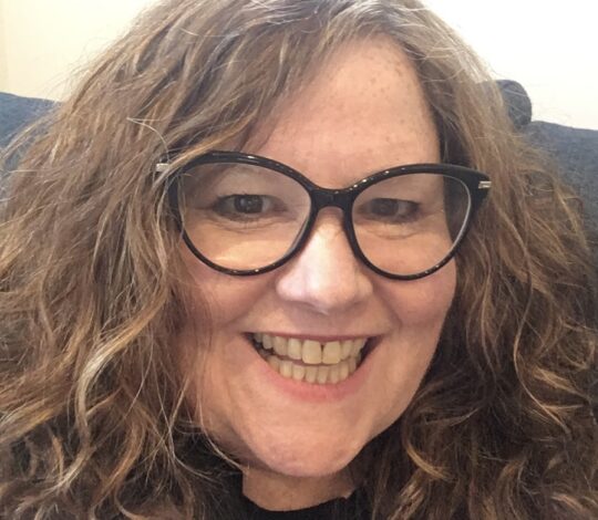 A person with long, curly brown hair and black glasses smiles broadly at the camera.