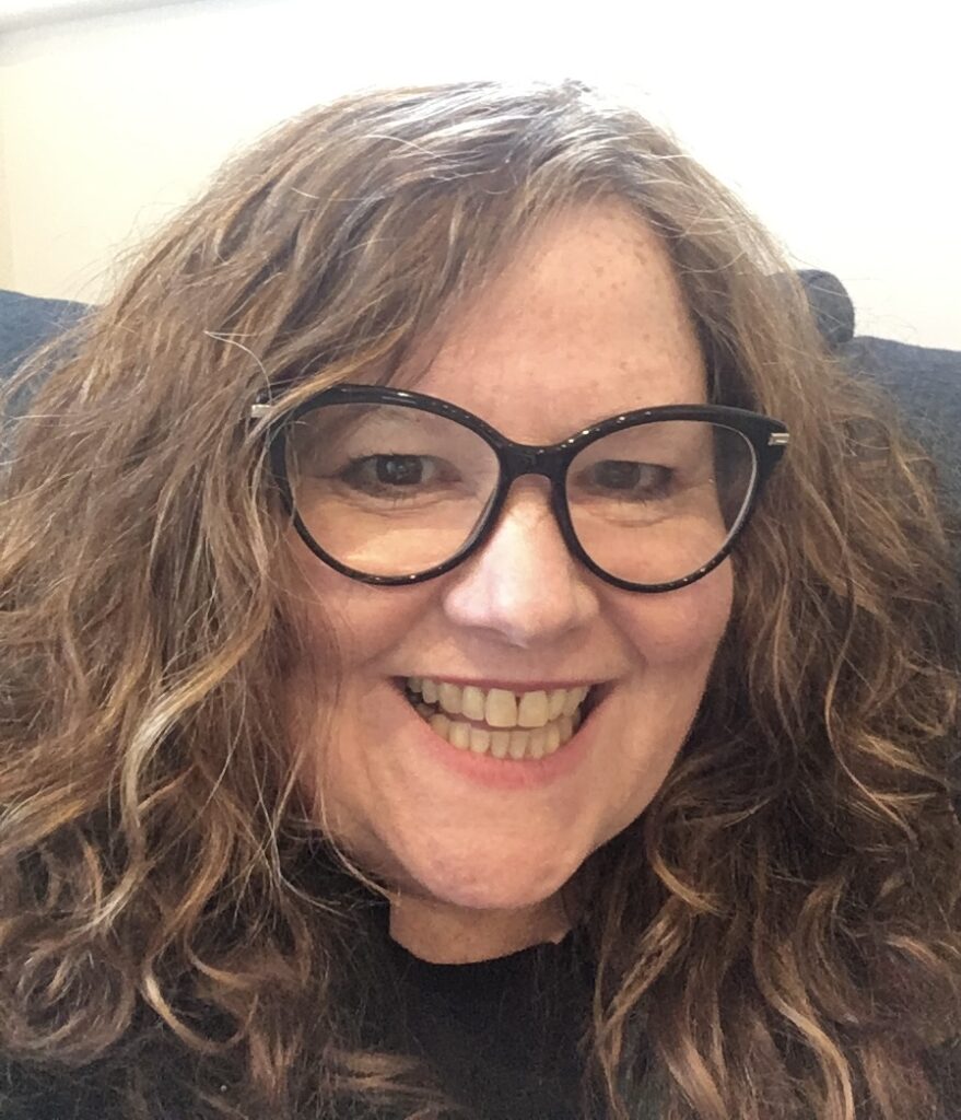 A person with long, curly brown hair and black glasses smiles broadly at the camera.