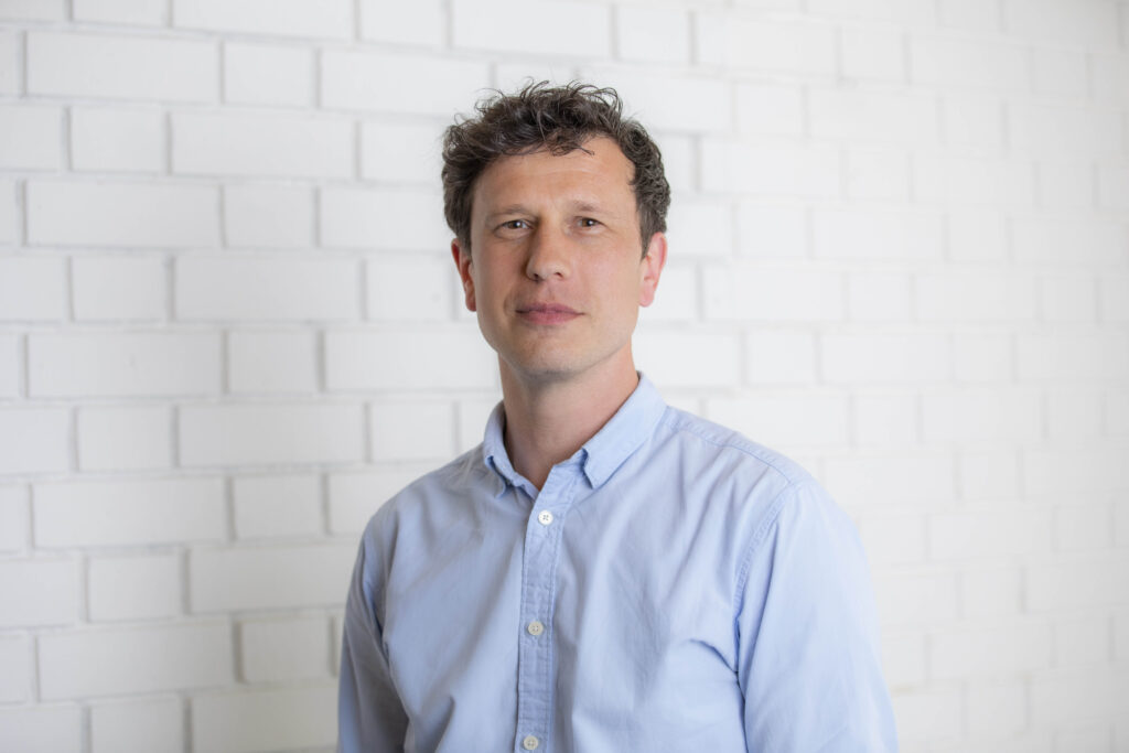 A man with short, curly hair, wearing a light blue button-down shirt, stands in front of a white brick wall.