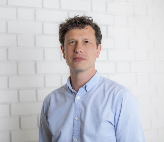 A man with short, curly hair, wearing a light blue button-down shirt, stands in front of a white brick wall.