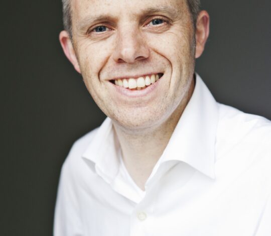 A person with short hair, wearing a white collared shirt, smiles at the camera against a dark background.