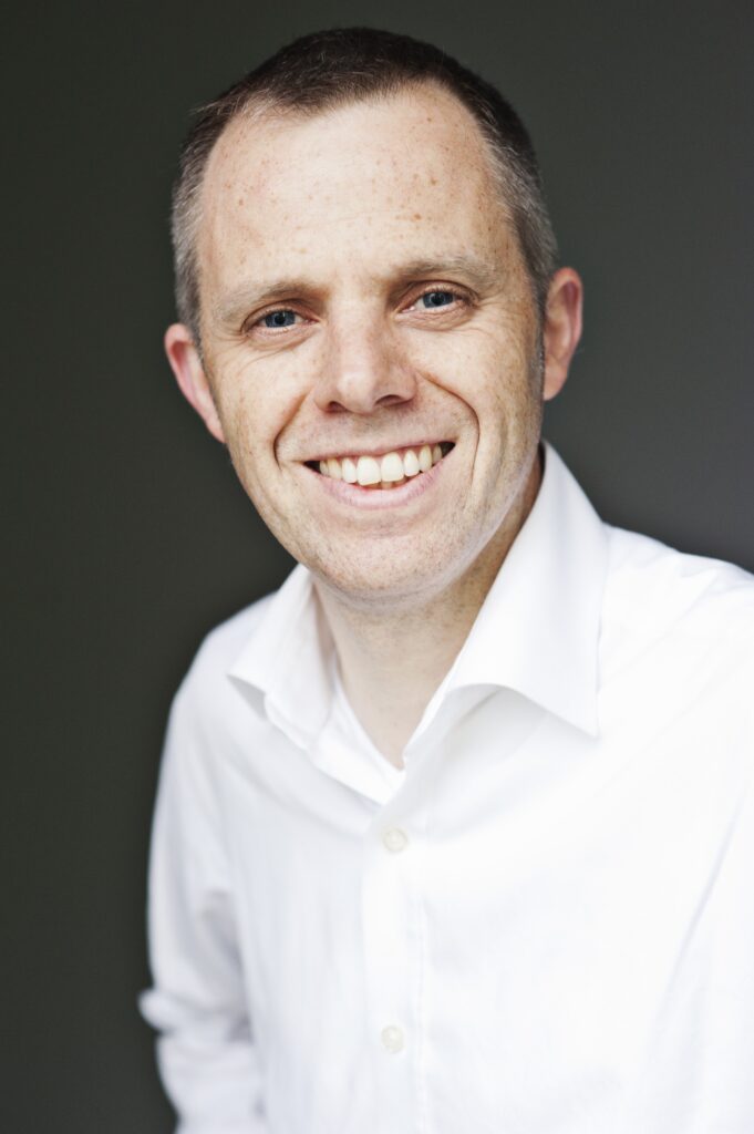 A person with short hair, wearing a white collared shirt, smiles at the camera against a dark background.