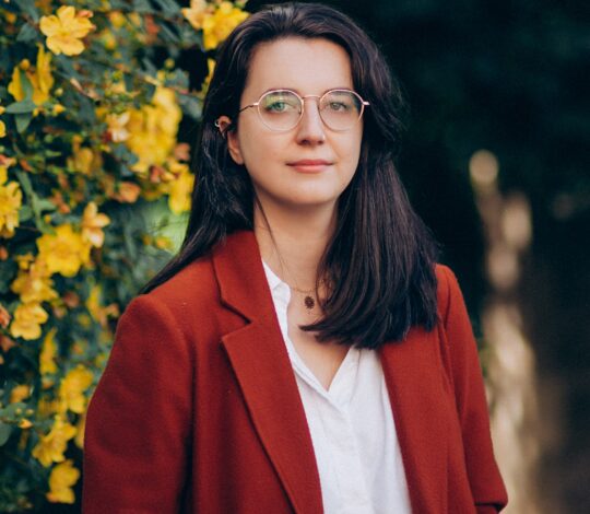 A person with long dark hair and glasses wearing a red coat and white shirt stands in front of yellow flowers and green foliage.
