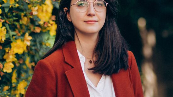 A person with long dark hair and glasses wearing a red coat and white shirt stands in front of yellow flowers and green foliage.