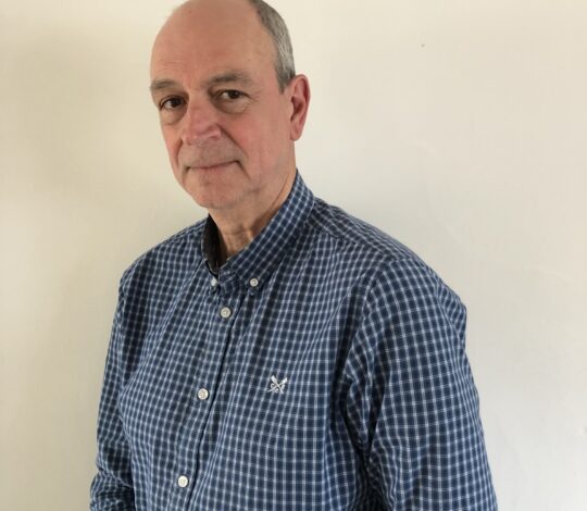 A middle-aged man with a receding hairline wearing a blue checkered shirt, standing against a plain white background.