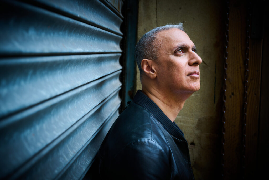 A man with short gray hair and a black jacket looks up while standing against a corrugated metal wall.