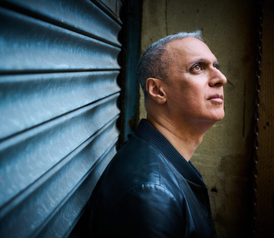 A man with short gray hair and a black jacket looks up while standing against a corrugated metal wall.