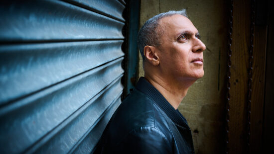 A man with short gray hair and a black jacket looks up while standing against a corrugated metal wall.