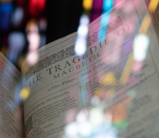 Open book showing the title "The Tragedie of Macbeth" with reflections of stained glass windows in the background.