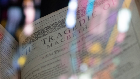 Open book showing the title "The Tragedie of Macbeth" with reflections of stained glass windows in the background.