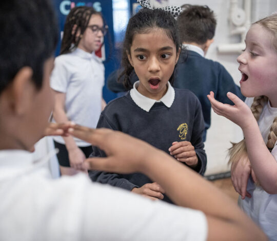 Three children stand indoors, one with an open mouth looking surprised, another gesturing with hands, and a third smiling and talking. Others are in the background. They all wear school uniforms.