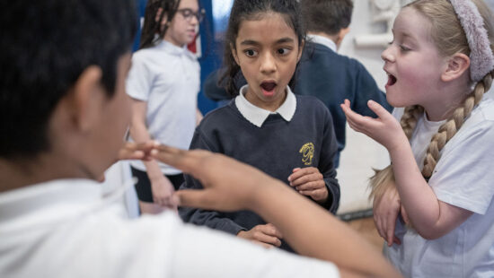 Three children stand indoors, one with an open mouth looking surprised, another gesturing with hands, and a third smiling and talking. Others are in the background. They all wear school uniforms.