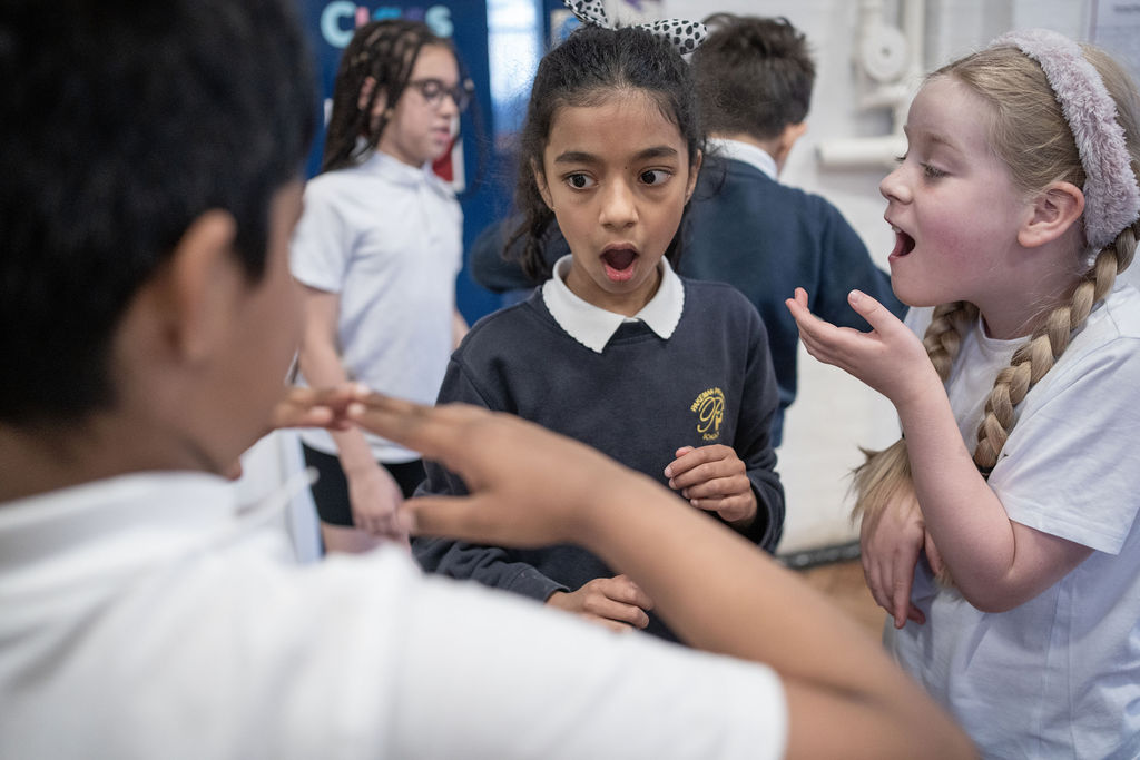 Three children stand indoors, one with an open mouth looking surprised, another gesturing with hands, and a third smiling and talking. Others are in the background. They all wear school uniforms.