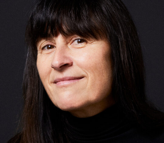 A woman with long dark hair and bangs, wearing a black turtleneck, smiles slightly against a dark background.