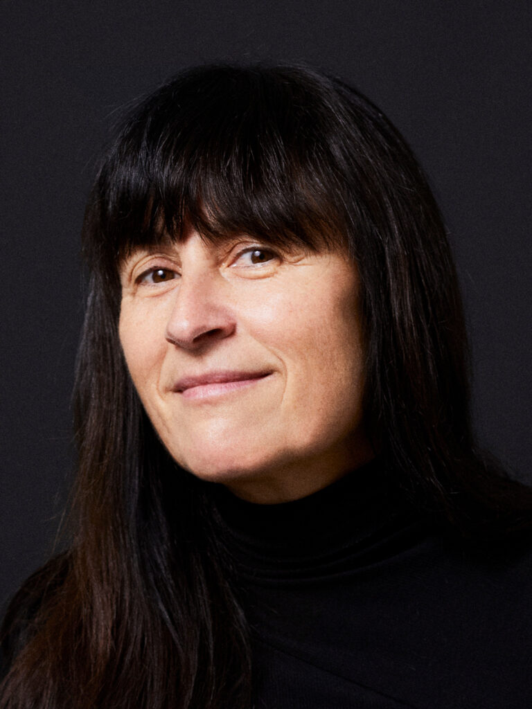 A woman with long dark hair and bangs, wearing a black turtleneck, smiles slightly against a dark background.