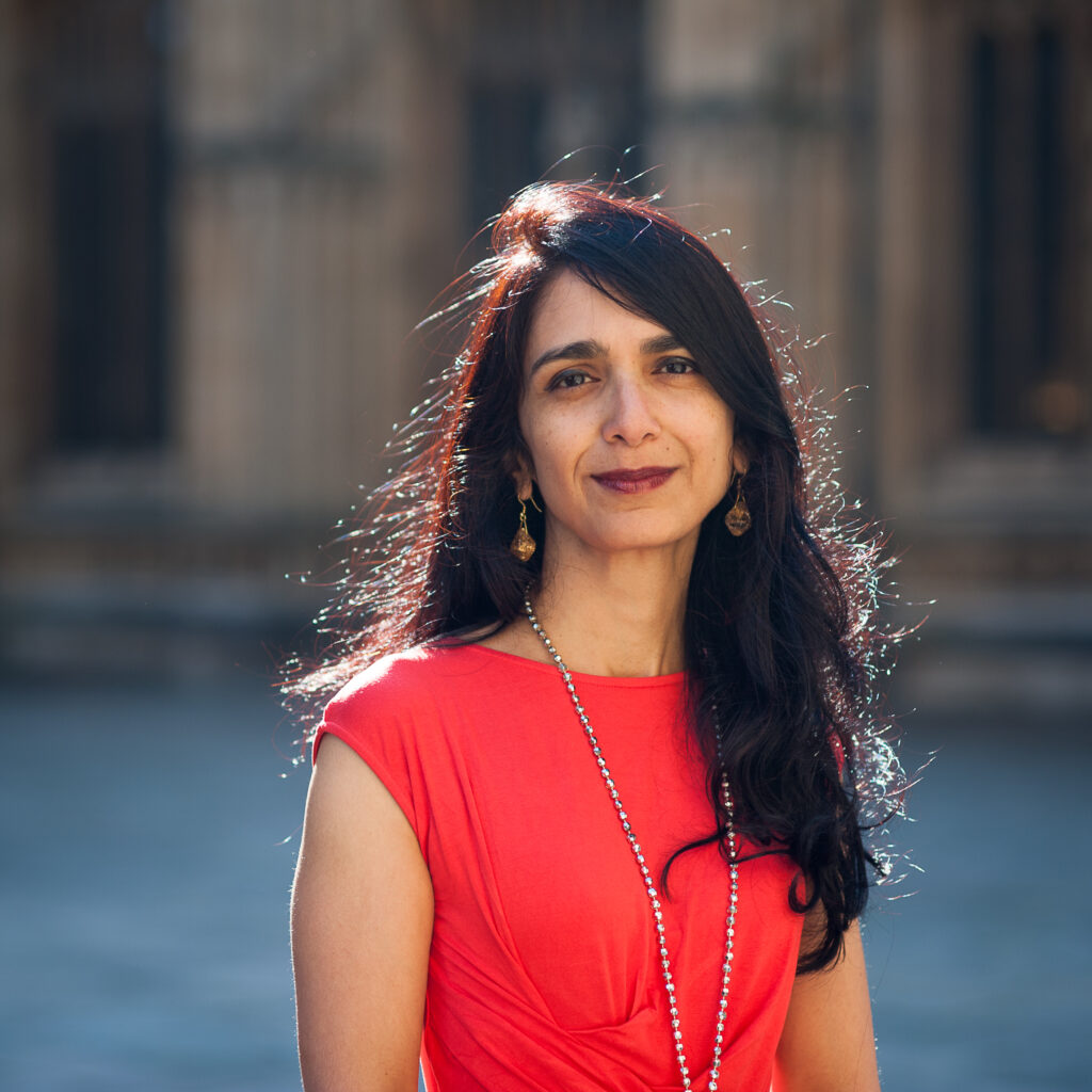 A woman with long dark hair wearing a red dress and a necklace stands outdoors, smiling slightly. The background is blurred.