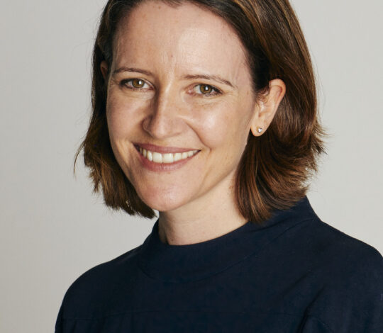 A woman with shoulder-length brown hair smiles at the camera against a plain background, wearing a dark blue top.