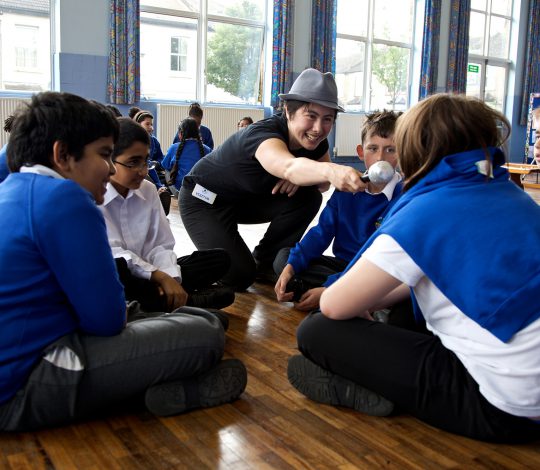 A group of schoolchildren sitting in a circle on a wooden floor, while an adult wearing a hat holds a microphone towards one of the children in a classroom setting with large windows.