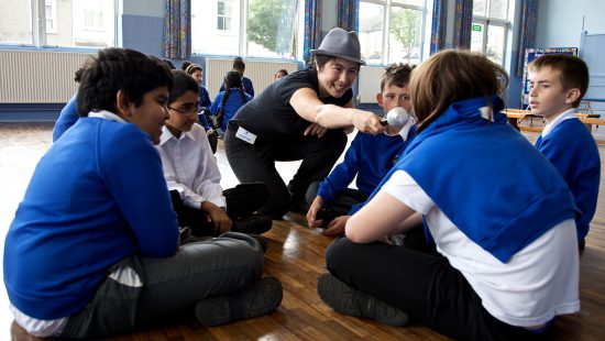 A group of schoolchildren sitting in a circle on a wooden floor, while an adult wearing a hat holds a microphone towards one of the children in a classroom setting with large windows.