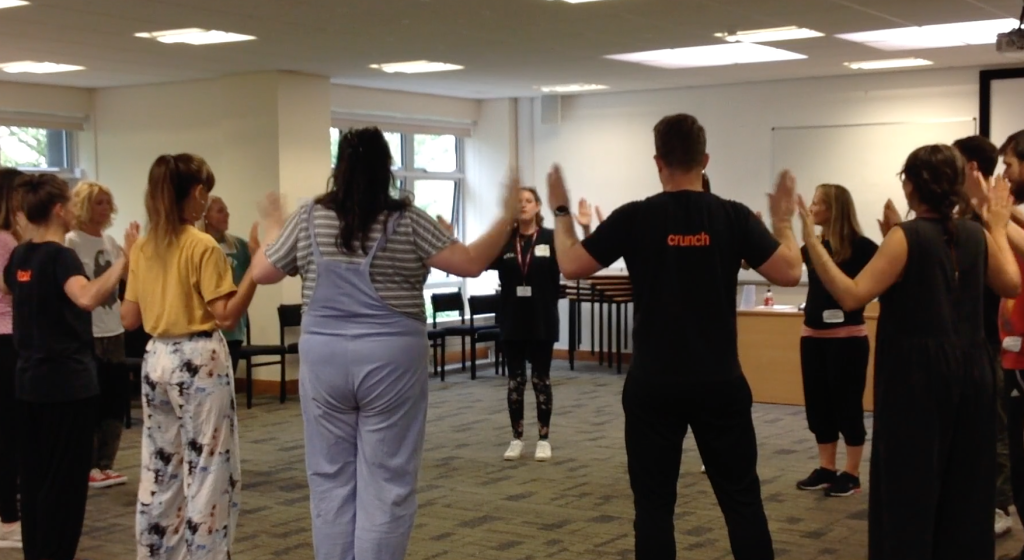 A group of people stand in a circle inside a room, raising their hands. One person appears to be leading the activity.