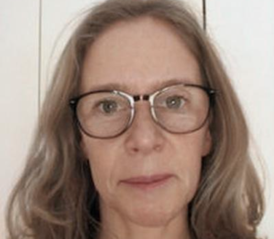 A woman with glasses and shoulder-length hair looks directly at the camera against a plain background.