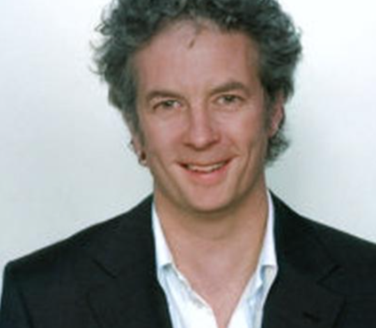 A man with curly gray hair is wearing a dark suit jacket over a white shirt, posing and smiling against a light-colored background.