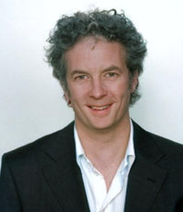 A man with curly gray hair is wearing a dark suit jacket over a white shirt, posing and smiling against a light-colored background.