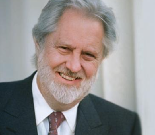 Elderly man with gray hair and beard, wearing a dark suit, white shirt, and patterned tie, smiles while standing in front of a white curtain.