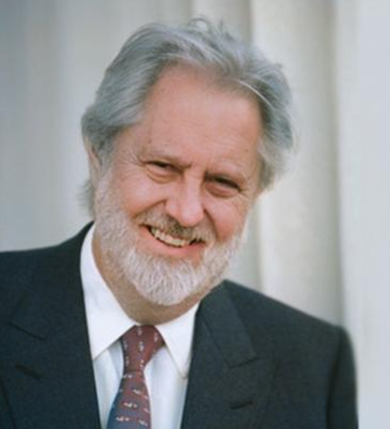 Elderly man with gray hair and beard, wearing a dark suit, white shirt, and patterned tie, smiles while standing in front of a white curtain.