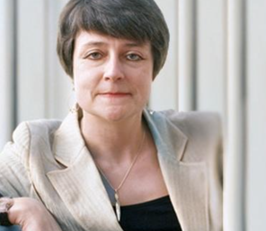 A woman with short dark hair wearing a beige blazer and a black top sits against a backdrop of vertical blinds.