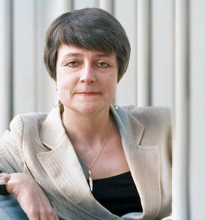 A woman with short dark hair wearing a beige blazer and a black top sits against a backdrop of vertical blinds.