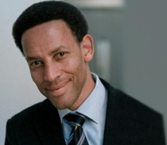 A person with short hair, wearing a suit and tie, smiles while facing the camera.