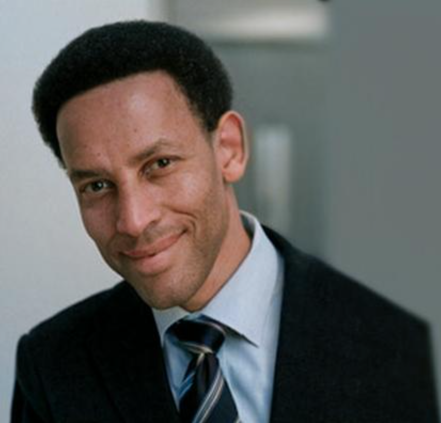 A person with short hair, wearing a suit and tie, smiles while facing the camera.