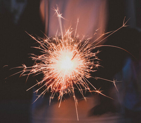 A close-up of a person holding a lit sparkler in the dark, emitting bright sparks and light. The person's silhouette and background are darkened.