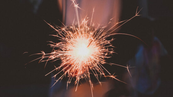 A close-up of a person holding a lit sparkler in the dark, emitting bright sparks and light. The person's silhouette and background are darkened.