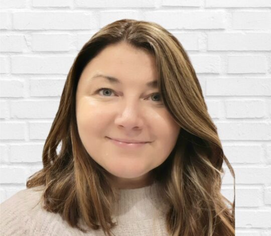 A woman with shoulder-length, light brown hair, wearing a light-colored sweater, stands in front of a white brick wall.