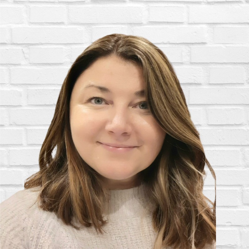 A woman with shoulder-length, light brown hair, wearing a light-colored sweater, stands in front of a white brick wall.