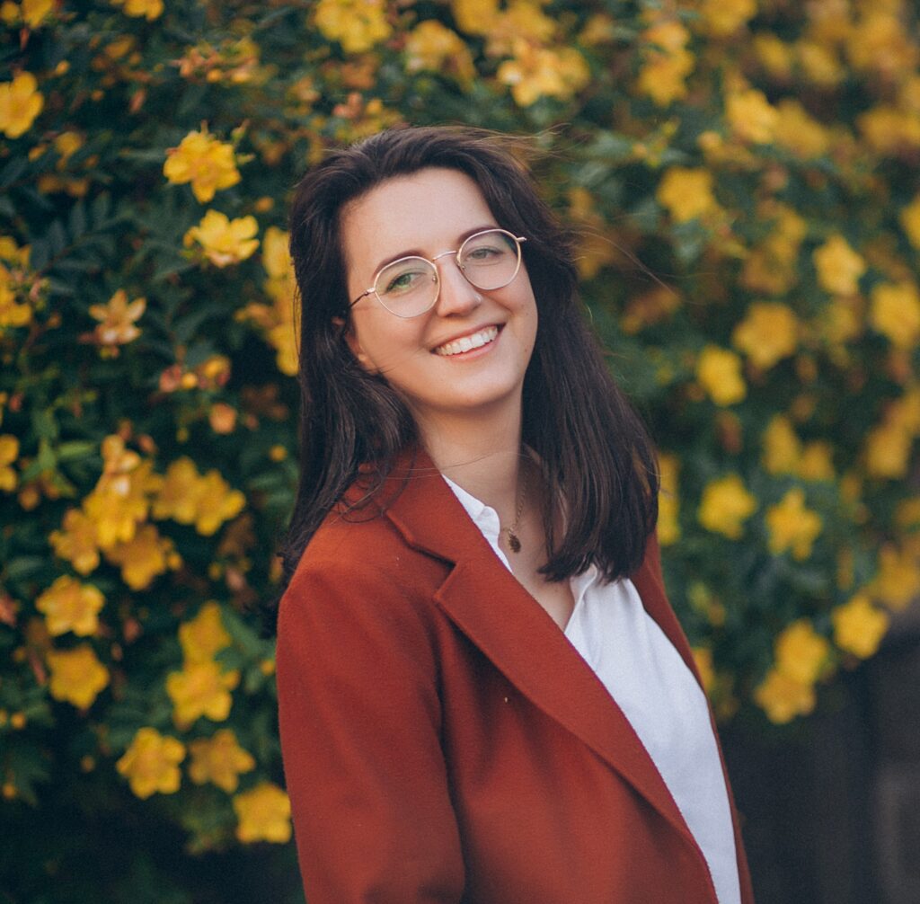 Woman with glasses and a red blazer smiling, standing in front of a bush with yellow flowers.