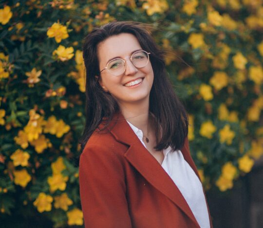 Woman with glasses and a red blazer smiling, standing in front of a bush with yellow flowers.