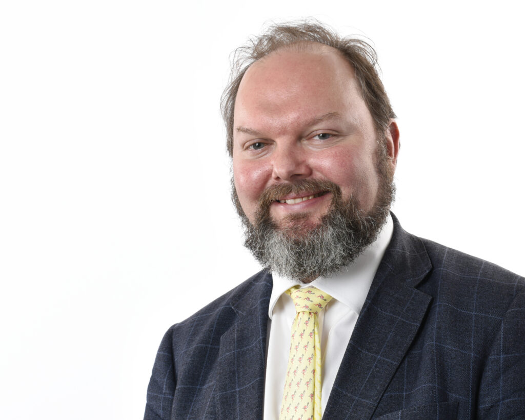 A bearded man in a suit with a white shirt and yellow tie is smiling against a plain white background.