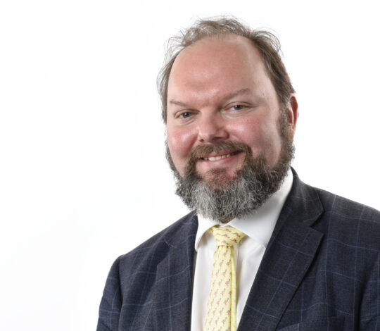 A bearded man in a suit with a white shirt and yellow tie is smiling against a plain white background.