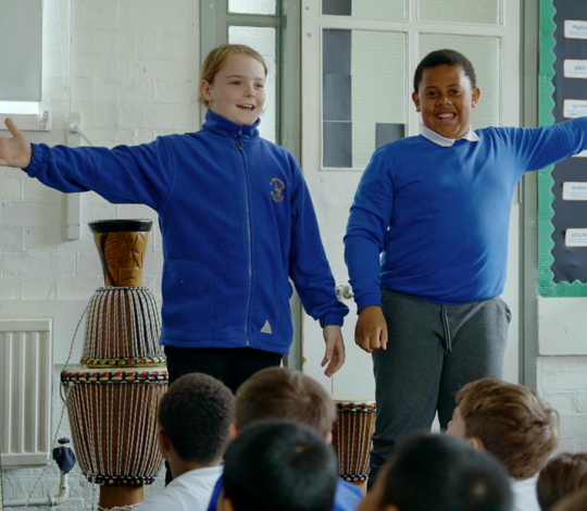 Two children in blue tops stand in front of an audience, arms outstretched, with a drum in the background. They appear to be performing in a classroom.