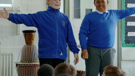 Two children in blue tops stand in front of an audience, arms outstretched, with a drum in the background. They appear to be performing in a classroom.