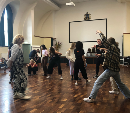 A group of people engaged in expressive movements in a large room with wooden floors, high ceilings, and a coat of arms on the wall.