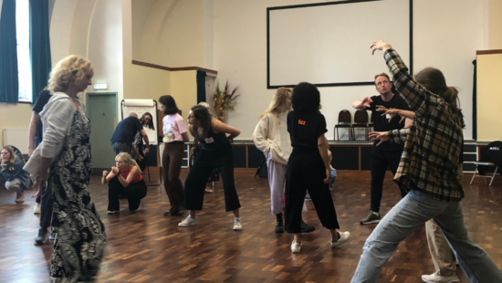 A group of people engaged in expressive movements in a large room with wooden floors, high ceilings, and a coat of arms on the wall.